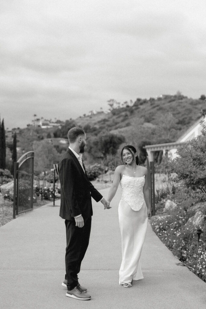 Kiana and Matt walking hand-in-hand and smiling at each other during their engagement session at Monserate Winery in Southern California, with vineyard hills in the background.