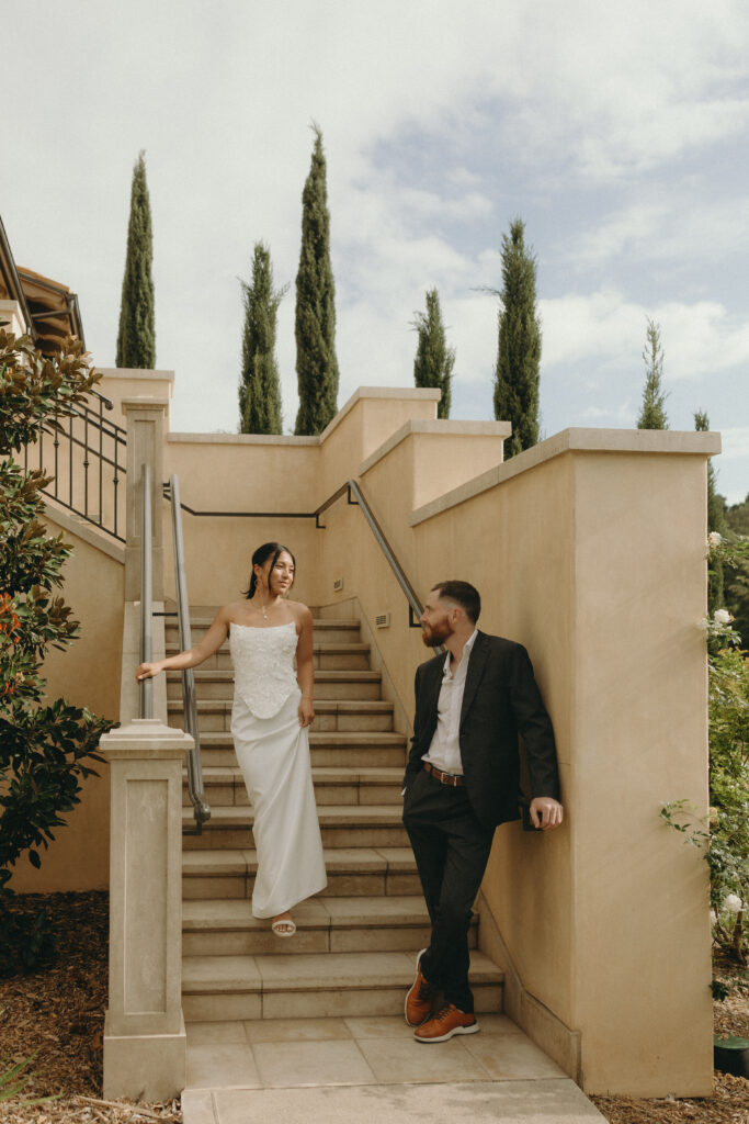 Kiana walking down stone steps toward Matt during their Monserate Winery engagement session, surrounded by Mediterranean-style architecture and tall cypress trees.