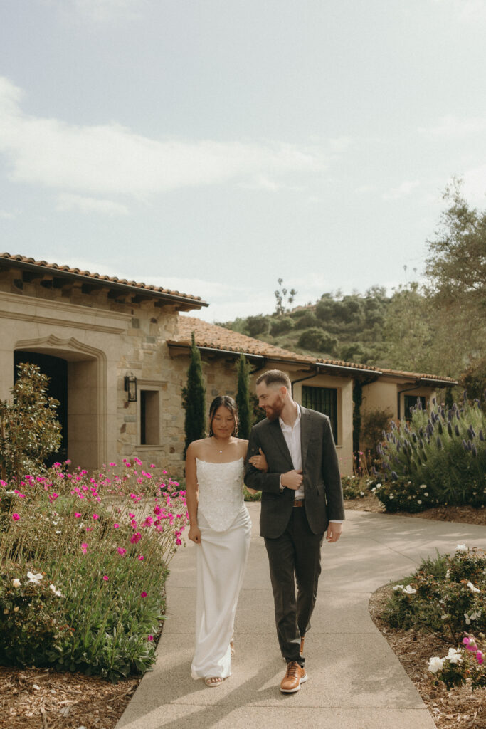 Kiana and Matt walking arm-in-arm through the flower gardens at Monserate Winery during their engagement session, surrounded by blooming pink and purple florals and Mediterranean-style architecture.